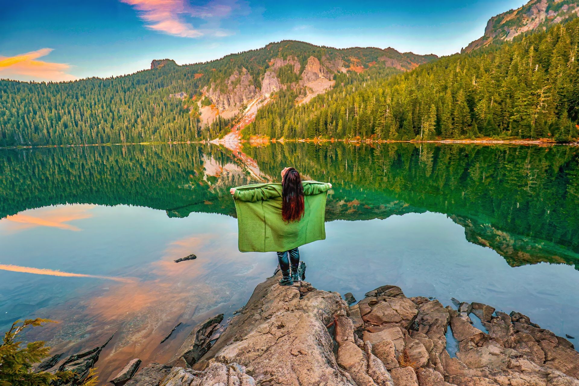 Person standing on rocks by a lake surrounded by forests and mountains, holding a green blanket open.