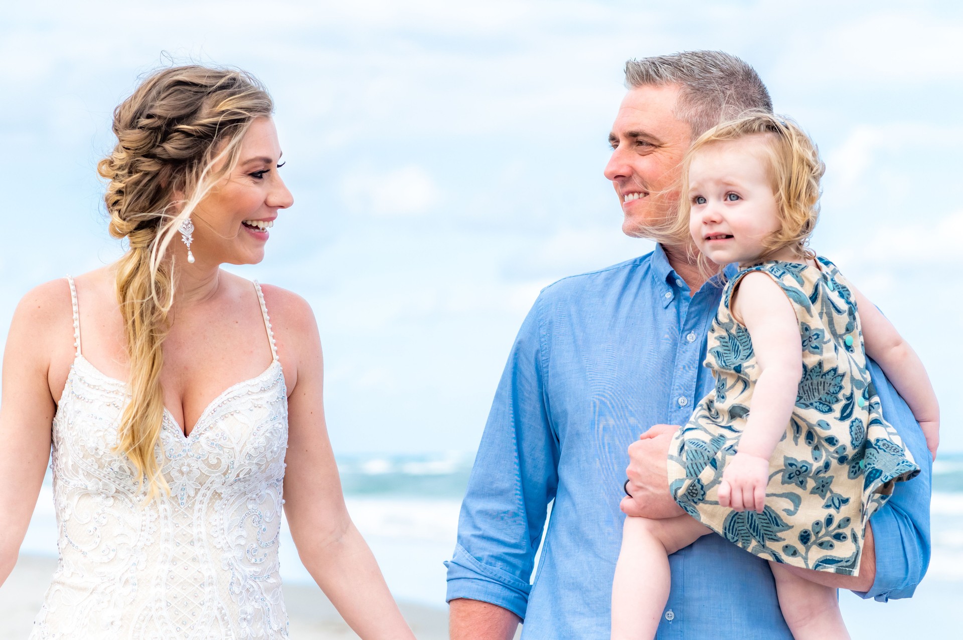 Beautiful bride holds a cute baby toddler on a beach in wedding dress. Little family destination wedding or elopement